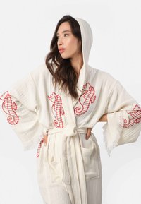 Krémový kapucňový plášť s vyšitým červeným morským koníkom, širokými rukávmi, zaväzovacím pásom a dvoma prednými vreckami. Textúrovaná tkanina s strapcovým lemom.