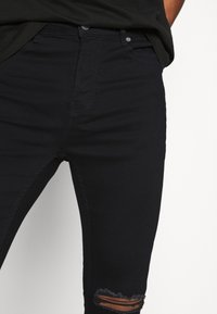 Jeans skinny neri con dettaglio strappato al ginocchio, realizzati in un tessuto elasticizzato con una superficie liscia e un classico design a cinque tasche.