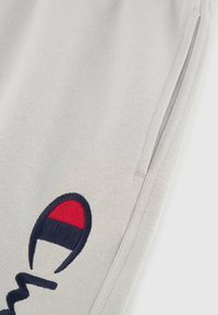 Pantalons de survêtement gris avec une poche latérale, arborant un logo brodé bleu marine et rouge. Le matériau est en coton doux avec une finition texturée.
