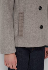 Manteau en mélange de laine gris clair avec deux boutons à l'avant, détails de poches texturées côtelées et coupe ajustée. Porté sur un jean en denim foncé.