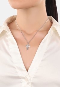 Collana d'argento a forma di croce con una superficie liscia e piatta, appesa a una catena sottile e delicata, indossata con una camicia chiara con colletto.