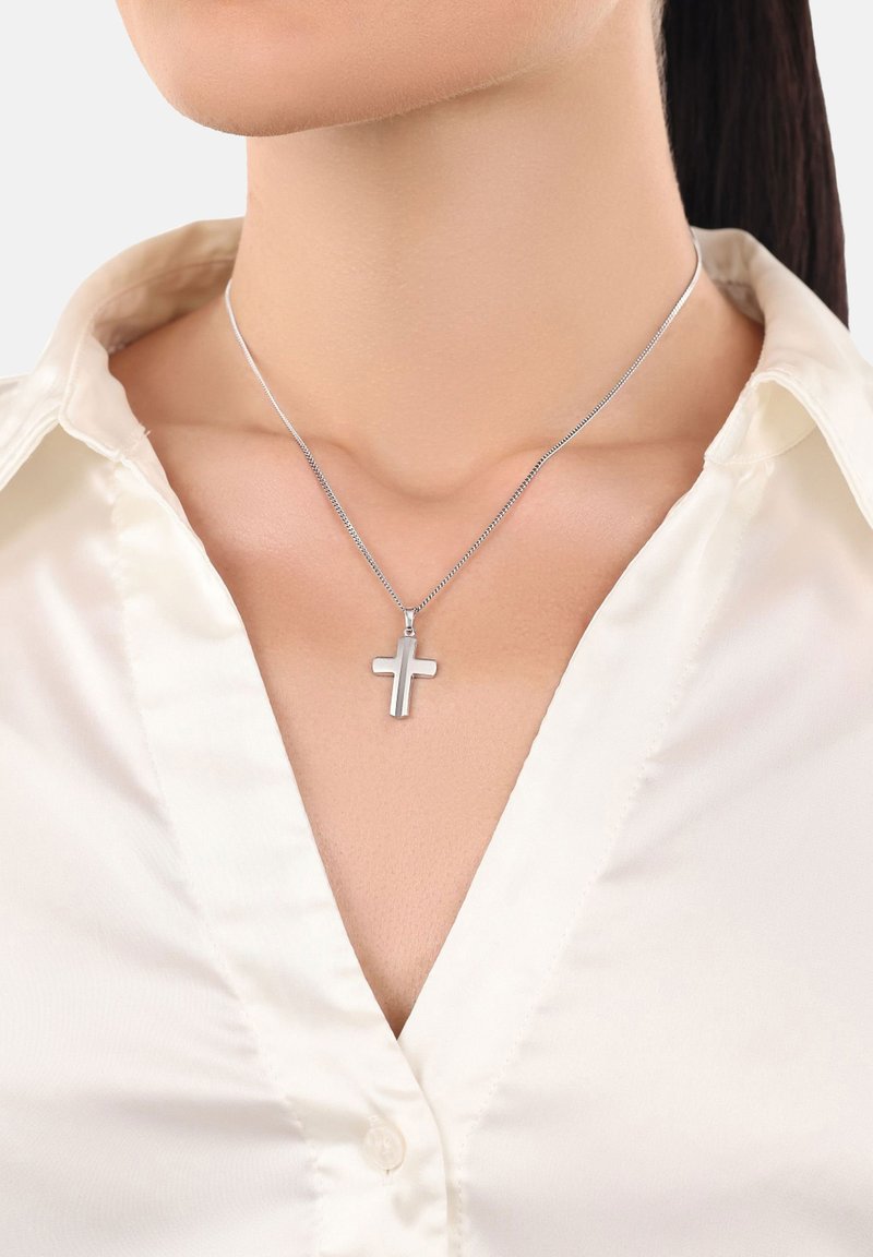 Collana d'argento a forma di croce con una superficie liscia e piatta, appesa a una catena sottile e delicata, indossata con una camicia chiara con colletto.