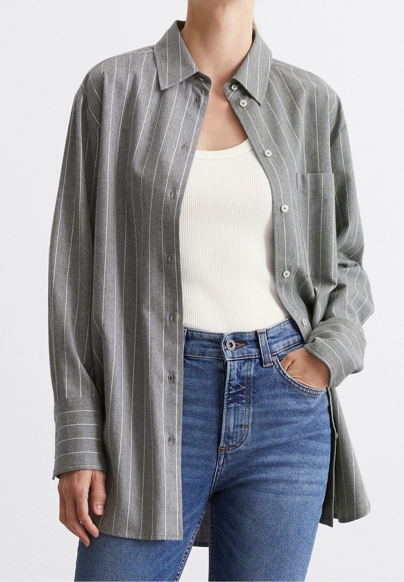 Personne portant un jean bleu, un débardeur blanc côtelé et une chemise grise ample à fines rayures et boutonnée, une main dans la poche.