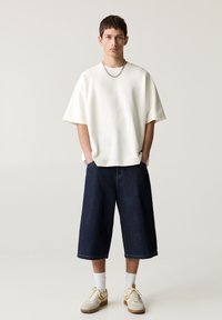 Chemise blanche oversize à manches courtes, culottes en denim noir, et baskets beige avec des accents jaunes. Un collier en chaîne simple ajoute du détail.