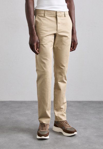 Chinos beiges en coton avec une coupe slim. Détails comprenant des poches latérales et une taille lisse. Associés à des chaussures de sport marron.