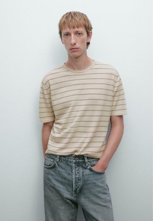 T-shirt a righe beige realizzata in tessuto morbido, con vestibilità rilassata e collo tondo, abbinata a jeans blu sbiaditi.