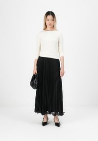 Top branco de mangas longas combinado com uma saia maxi plissada preta, em tecido suave, e uma bolsa de mão preta, realçado por sapatos pretos de bico.