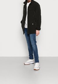 Chaqueta negra esencial con múltiples bolsillos, cierre de botones y un cuello alto, combinada con jeans azules y zapatillas blancas.