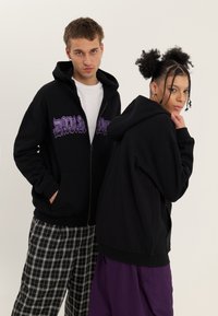 Jeune homme et femme portant des sweats à capuche noirs ; l'homme en pantalon à carreaux avec les mains dans les poches, la femme en pantalon violet avec la main près du menton.