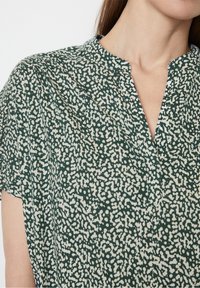 Blusa verde a fantasia con scollo a V, maniche corte e un tessuto morbido e testurizzato con macchioline bianche.