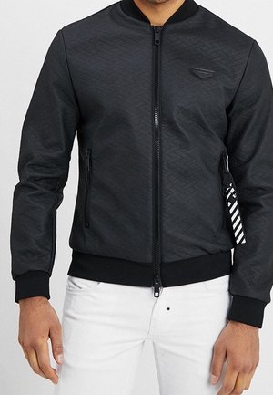 Mann trägt eine schwarze Bomberjacke mit Muster und Reißverschlusstaschen sowie weiße Hose, Hände entspannt an den Seiten.