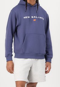 Lila hoodie med en framficka, med "NEW BALANCE" i vit och orange text. Matchad med ljusgrå shorts. Tillverkas i bomull.