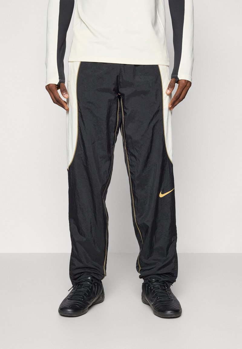 Schwarze Windbreaker-Hose mit goldenen Akzenten und Nike-Logo, ausgestattet mit einer leichten, geknitterten Textur und kontrastierenden weißen Seitenpaneelen.