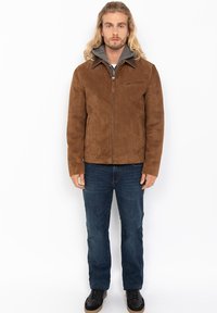 Uomo con lunghi capelli biondi che indossa una giacca di suede marrone, un felpa grigia, jeans blu e sneaker nere, in piedi contro uno sfondo bianco.