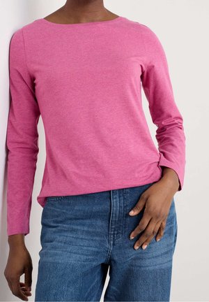 Osoba ubrana w różową koszulę z długim rękawem i niebieskie dżinsy, stojąca z jedną ręką opartą na biodrze na neutralnym tle.