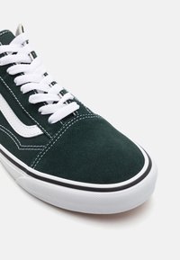 Gros plan d'une basket vert foncé avec des lacets blancs, une bande latérale blanche, une semelle en caoutchouc blanche et des coutures visibles sur l'orteil et les côtés.