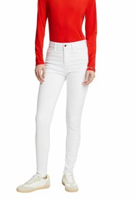 Jeans skinny blancos de denim, con una cintura alta y un botón de cobre. Combinados con una blusa roja de manga larga y zapatillas blancas.
