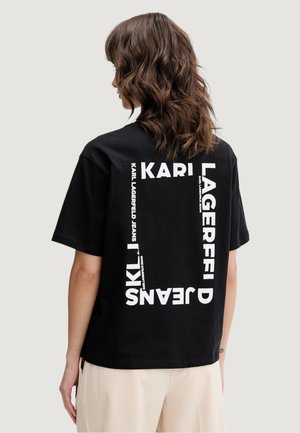 Kobieta o falowanych brązowych włosach ubrana w czarną koszulkę z białym napisem „KARL LAGERFELD JEANS” ułożonym w kwadrat na plecach.