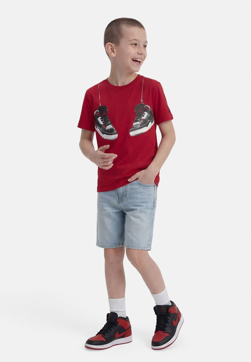 Smiling boy wearing a red T-shirt with printed sneakers, light denim shorts, white socks, and black-red high-top sneakers.