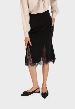 Femme portant une jupe midi noire à ourlet en dentelle et un chemisier beige à manches longues, assortis à des ballerines noires pointues avec bride arrière.