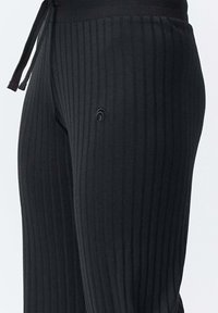 Nahaufnahme von schwarzen, gerippten Strickhosen mit Kordelzugbund und kleinem, rundem, gesticktem Logo am Oberschenkel.