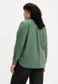 Camisa verde de manga larga hecha de una tela suave con cuello y puños abotonados, que presenta un sutil pliegue en la parte posterior y un dobladillo redondeado.