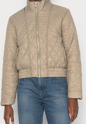 Beige quilted jacket with full front zipper, high collar, elastic waistband, worn with blue denim jeans.