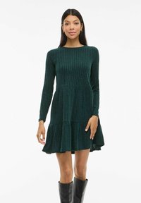 Femme portant une robe à manches longues en rib stop vert foncé avec un ourlet évasé, associée à des bottes noires jusqu'aux genoux, se tenant devant un fond blanc.