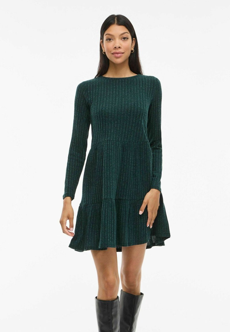 Femme portant une robe à manches longues en rib stop vert foncé avec un ourlet évasé, associée à des bottes noires jusqu'aux genoux, se tenant devant un fond blanc.