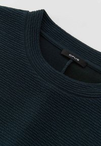 Dunkelgrünes strukturiertes Oberteil mit einem gerippten Design, ausgestattet mit einem Rundhalsausschnitt und einem schwarzen Etikett mit der Aufschrift "OPUS" auf der Innenseite.