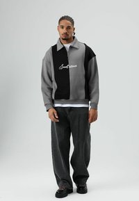 Grijs-zwarte sweatvest met rits en kraag, met witte geborduurde tekst, gecombineerd met losse zwarte jeans en zwarte schoenen.