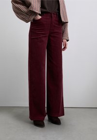 Pantalones de pana de pierna ancha en un profundo tono burdeos, con cintura alta, bolsillos frontales y textura suave, combinados con una chaqueta de cuadros.