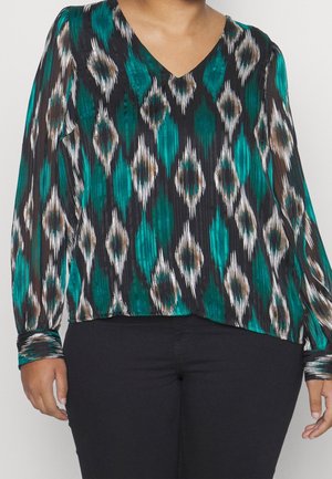 Woman wearing a long-sleeve, V-neck blouse with teal, black, white, and brown diamond pattern, paired with black pants.