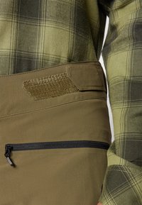 Pantalons vert olive avec une poche zippée noire et une patch en Velcro texturé. Associés à une chemise en plaid vert et noir.