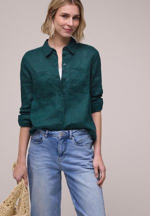 Mujer rubia con camisa verde oscuro con botones remangada y vaqueros azul claro, sosteniendo un bolso tejido beige en la mano izquierda.