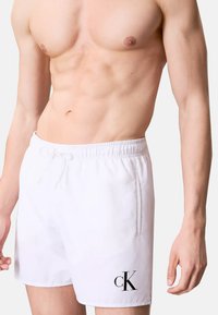 Pantaloni da bagno bianchi con vita elasticizzata e coulisse. Presentano un logo "cK" nero sul lato sinistro in basso. Tessuto liscio, vestibilità normale.