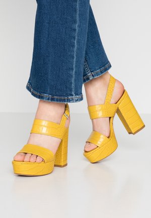 Pieds portant des escarpins à plateforme jaune vif, à motif crocodile, avec des brides autour de la cheville, assortis à un jean bleu denim court.