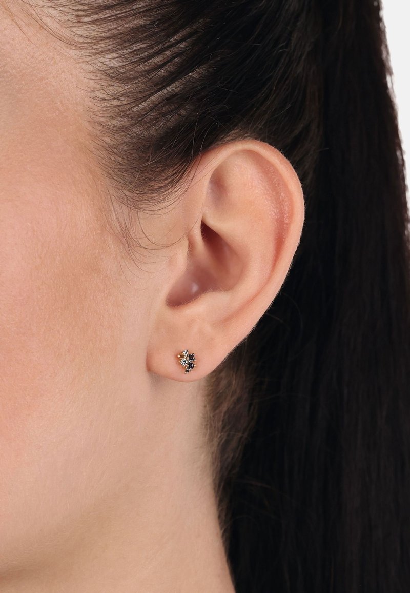 Gros plan sur l'oreille d'une femme aux cheveux foncés tirés en arrière, portant une petite boucle d'oreille clou noire et dorée sur le lobe de l'oreille.