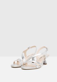 Sandales ouvertes en métal argenté avec des lanières courbées, un petit talon bloc et une boucle réglable à la cheville. Texture lisse avec un design minimaliste.