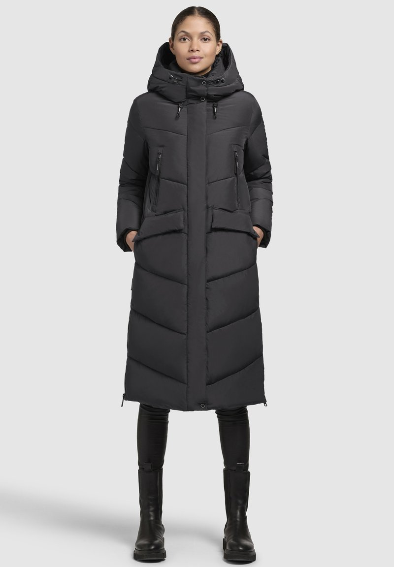 Černý dlouhý péřový kabát s kapucí, s diagonálně prošívaným vzorem, dvěma zipovými kapsami a rovný střih. Nosený přes černé kalhoty a boty.