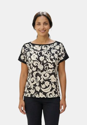 Donna con capelli scuri raccolti in una coda bassa, che indossa una maglietta a maniche corte bianca e nera con motivo floreale e arabeschi.