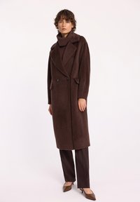 Długi brązowy wełniany płaszcz z wykładanym kołnierzem, podwójnymi guzikami i dużymi kieszeniami, połączony z ciemnobrązowymi spodniami i spiczastymi butami.