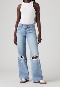 Světle modré džíny s širokými nohavicemi a opranými detaily na kolenou, s designem pěti kapes a kovovými knoflíky, doplněné bílým nátělníkem.