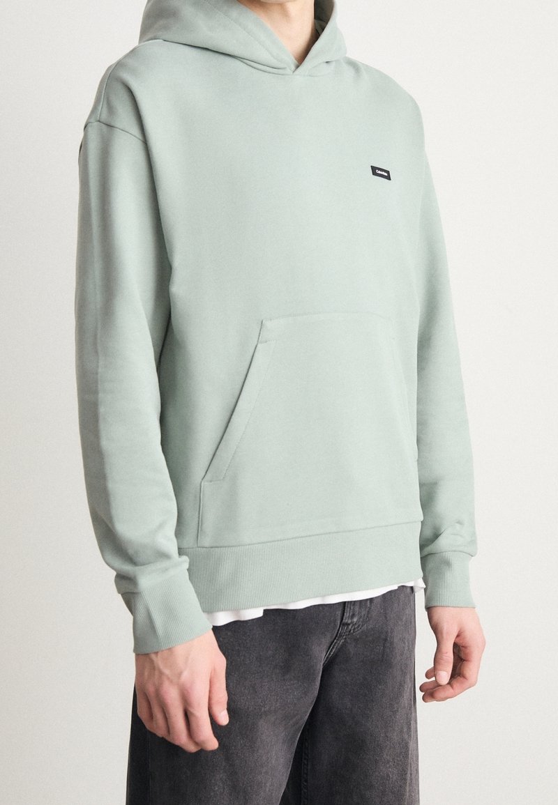 Sudadera con capucha en verde claro, de tela de mezcla de algodón. Presenta un bolsillo frontal y puños acanalados, con una pequeña etiqueta de marca negra en el pecho.