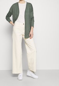 Zielony długi kardigan z dzianiny noszony na białej koszuli, zestawiony z kremowymi spodniami o szerokich nogawkach i białymi sportowymi butami. Kardigan ma otwarty przód.