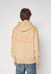 Sudadera beige con diseño de contorno naranja en la parte posterior, con un bolsillo tipo canguro y puños y dobladillo acanalados. Combinada con unos jeans azules.