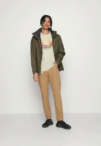 Zelena jakna sivo notranjostjo, kremast majica z rdečo in oranžno grafiko gore, svetlo rjave hlače in črne čevlje.