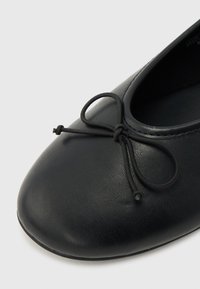 Μαύρο δερμάτινο μπαλαρέτα με στρογγυλεμένη μύτη και διακοσμητικό δεμένο φιόγκο στο επάνω μέρος, εμφανίζεται πάνω σε ανοιχτό γκρι φόντο.