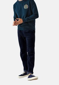 Felpa in cotone blu navy con logo ricamato, abbinata a pantaloni di corduroy scuri e sneakers blu con suola e lacci crema.