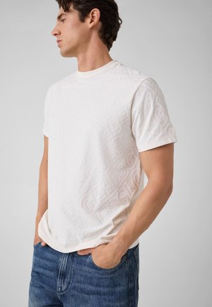 Homme debout, main gauche dans la poche de jean, portant un t-shirt blanc embossé avec un motif de texte subtil, regardant vers la gauche.